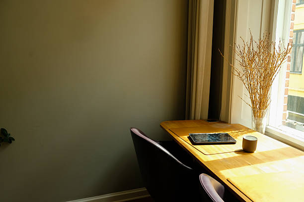 A clean and minimalist home office workspace featuring an open laptop, a ceramic mug, and a vase of dried branches placed on a wooden desk beside a large sunlit window. This warm, modern scene represents remote work, digital productivity, flexible work culture, and contemporary workspace design. Ideal for concepts such as freelancing, creative work, hybrid work environments, focus time, and digital nomad lifestyle.