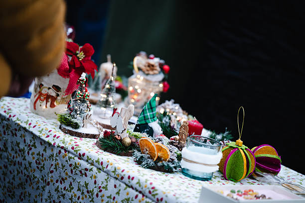 A festive assortment of handmade Christmas decorations displayed on a patterned market tablecloth. Miniature trees, ornaments, candles, dried oranges, and colorful crafts create a cozy holiday atmosphere. Soft lighting highlights the textures and vibrant seasonal details.