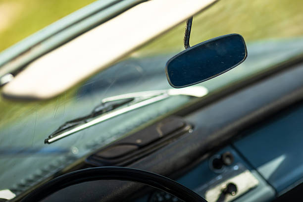 Close-up of a vintage car interior focusing on the rearview mirror, sun visor, and dashboard seen through the windshield in natural daylight. The image highlights classic automotive design, simple interior features, and a nostalgic driving atmosphere, ideal for themes about retro cars, safety, and travel.