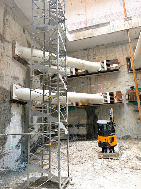 Vertical view showing massive steel shoring struts connected to a concrete diaphragm wall at the Grand Paris Express construction site with a yellow excavator below.