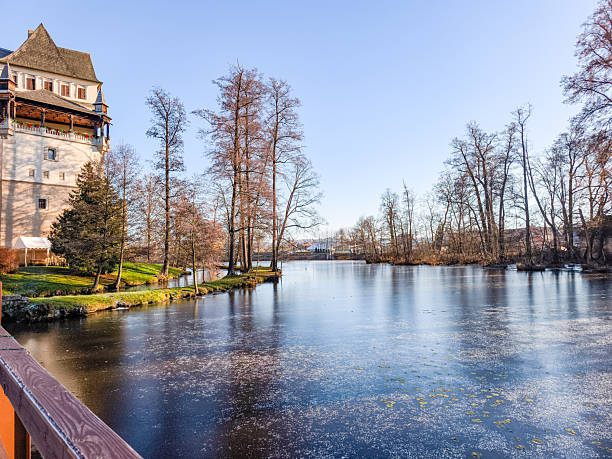 Blatna, Czech republic - November 23, 2025: Blatna castle on a sunny winter day, reflecting on the partially frozen pond with bare trees contrasting a clear blue sky
