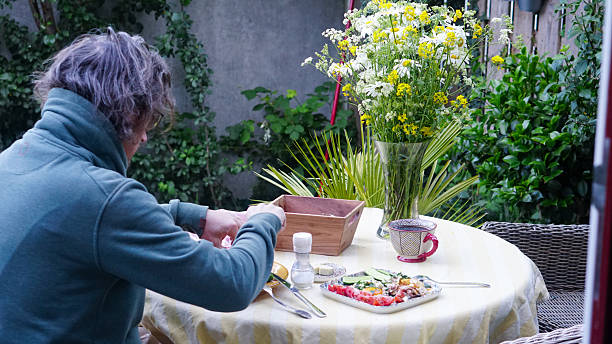 A man with curly hair seated at a round table outdoors, preparing or eating a meal in a garden setting. The table is covered with a light yellow cloth and set with a plate of food, a patterned cup, a salt shaker, butter dish, and a wooden box. A large glass vase with white and yellow wildflowers adds freshness and visual warmth. Surrounded by lush greenery and a quiet wall, the scene evokes balance, connection to nature, and the beauty of everyday rituals.