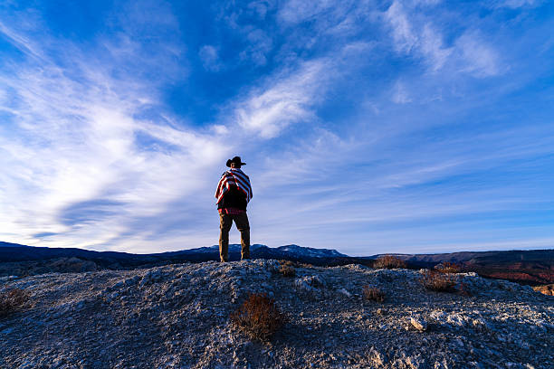 Patriotic American man with US flag draped over shoulders while looking off into the distance with scenic mountain views in background.
