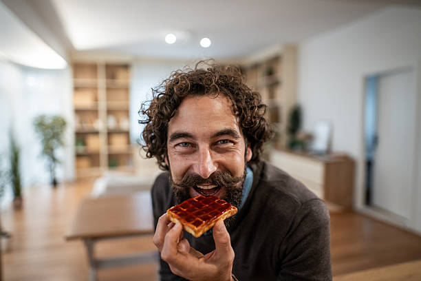 Smiling adult man with curly hair and mustache enjoying a sweet waffle with jam for breakfast, looking directly at the camera in a bright home setting