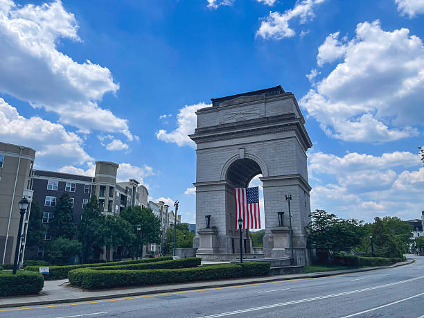 Majestic stone monument featuring a large arch and a hanging American flag, surrounded by greenery and modern residential buildings beneath a bright summer sky with dramatic clouds.