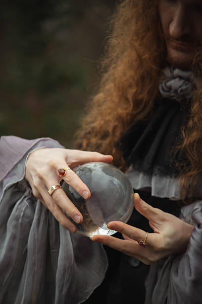A fortune teller with long curly hair holds a crystal ball in both hands. Dressed in an elegant outfit, they seem focused and contemplative while surrounded by nature.