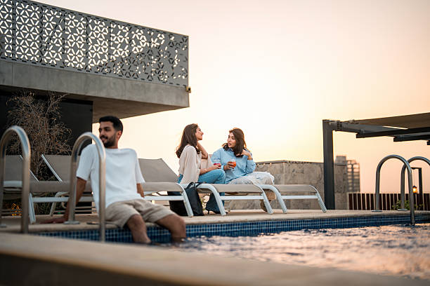 Low-angle view of young Middle Eastern adults in casual attire lounging on white deck chairs beside an elegant rooftop swimming pool, illuminated by warm sunset light.