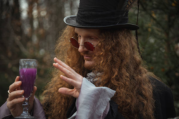 A man with long curly hair and a top hat stands in a forest during autumn. He wears sunglasses and examines a purple drink in a clear glass, creating a mysterious atmosphere.
