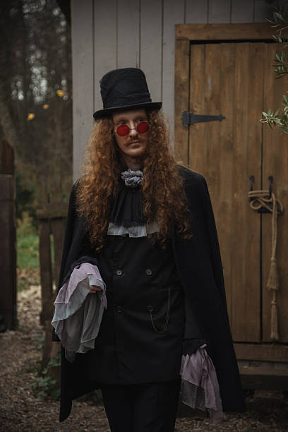 A person with long curly hair wears a vintage outfit, featuring a hat and round sunglasses. They walk confidently along a forest path near a wooden structure, surrounded by nature.