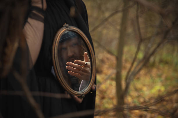 A shadowy figure stands in a forest filled with fall foliage, holding a vintage mirror. The reflection shows a hand reaching out, creating an eerie atmosphere in the woods.