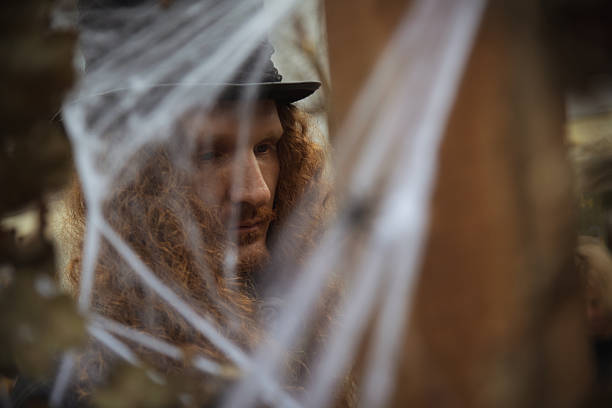 A man with long curly hair wears a hat and gazes thoughtfully in a spooky setting. Cobwebs and eerie decorations create a haunting atmosphere during a Halloween event.