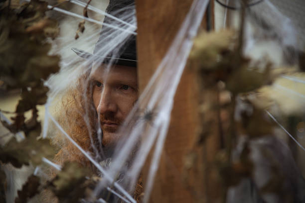 A man with curly hair and a top hat peers through a web-covered wooden structure. The atmosphere feels eerie and filled with suspense, suggesting a Halloween or haunted event.