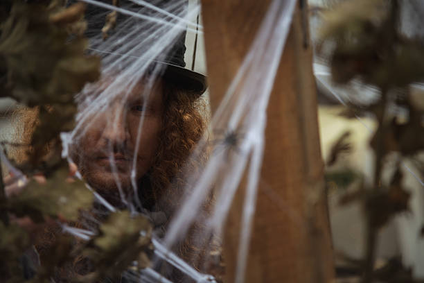 A figure with curly hair and a hat peers through a web-covered wooden structure, creating an eerie atmosphere. The scene is likely set for a Halloween event or haunted attraction.