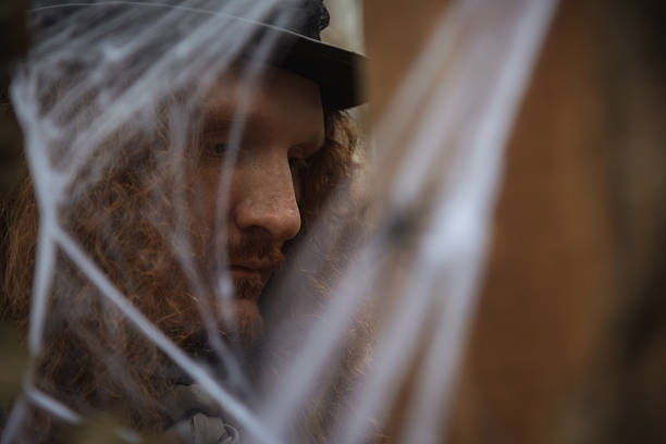 A man with curly red hair is thoughtfully gazing among Halloween decorations. He wears a hat and is surrounded by cobwebs, creating an eerie atmosphere at a festive event.