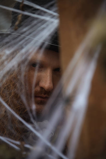 A man with a long beard is seen peeking through delicate spider webs, creating a mysterious atmosphere. The setting is dimly lit, enhancing the sense of intrigue and suspense.
