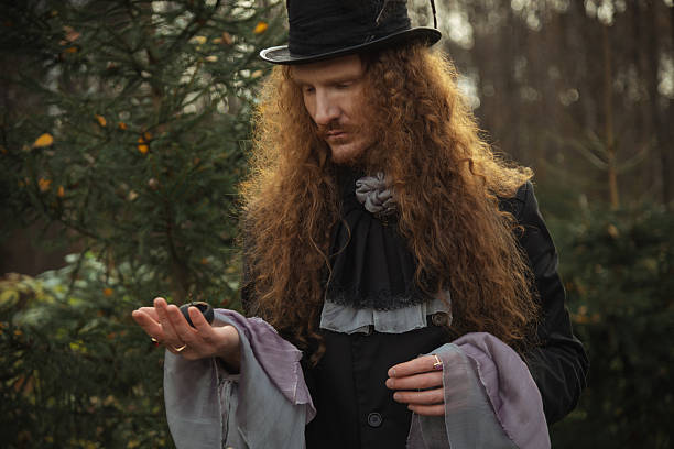 A man with long curly hair and a top hat stands in a peaceful forest, gently holding a small animal in his hand while wearing a vintage outfit with flowing sleeves.