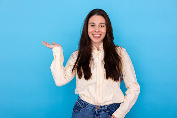 istock Young woman smiling, presenting concept or product with open palm on blue background, offering copy space 2249593584