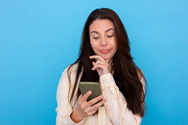 istock Young woman holding a smartphone, looking at its screen with a thoughtful expression. She is considering information or a decision 2249594458