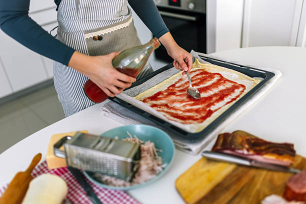 Young woman carefully pouring tomato sauce over pizza dough at home, preparing for toppings and baking.