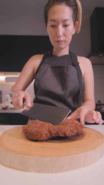 Asian woman cutting crispy breaded pork cutlet on wooden board, vertical