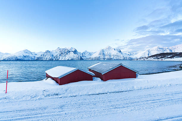 Dusk over wooden cabins by the fjord in the snowy landscape with rocky mountains covered with snow in the background, Lyngen Alps, Troms, Norway, Scandinavia