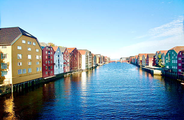 The image displays the historic colorful wooden buildings lining the Nidelva River in the Bakklandet district of Trondheim, Norway. This view is typically captured from the Gamle Bybro (Old Town Bridge).  ,  ,Norway - September 30,2025 Colorful buildings stand along the riverbank, reflecting in the clear water beneath a bright blue sky. The scene captures a serene moment in a vibrant urban setting.