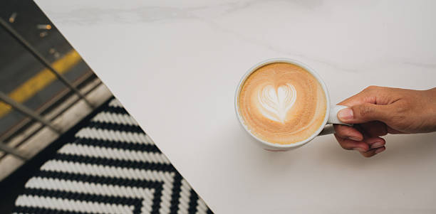 Latte coffee cup with heart shaped foam art held by hand on white table with black and white patterned rug in background, cozy and warm atmosphere