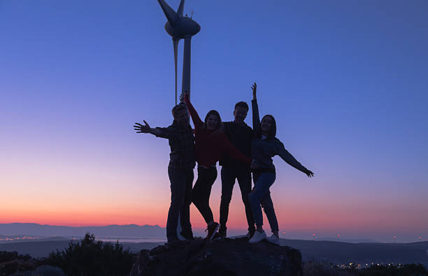 A group of four friends stands on a hilltop at a wind farm during blue hour, silhouetted against the colorful evening sky with open arms in a joyful, carefree pose. A large wind turbine rises in the background, connecting the themes of happiness, togetherness, and outdoor adventure with renewable energy, environmental awareness, and sustainability. This vibrant scene highlights the beauty of friendship, the excitement of exploring nature, and the promise of a cleaner, greener future
