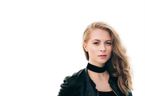 Close-up portrait of a young woman wearing a black leather jacket and choker against a bright white background. Soft natural light highlights her facial features and wavy blonde hair, creating a clean and modern aesthetic. The image conveys confidence, style, and contemporary fashion.
