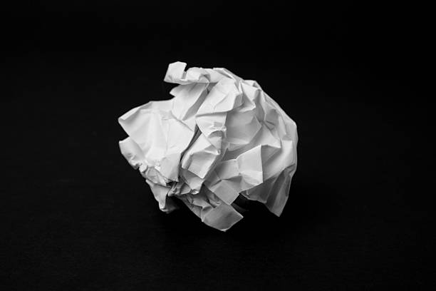 A single white crumpled paper ball sits on a dark black surface, highlighted by soft lighting that accentuates its folds and texture. The stark contrast creates a dramatic, minimalist composition. The image conveys concepts of discarded ideas, frustration, creativity, and simplicity.