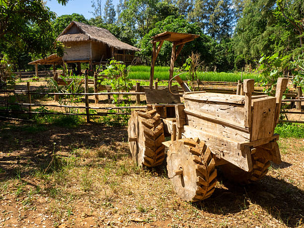A handcrafted wooden tractor standing before a raised thatched house, surrounded by fields and rural greenery. Reflects traditional Thai agricultural lifestyle.