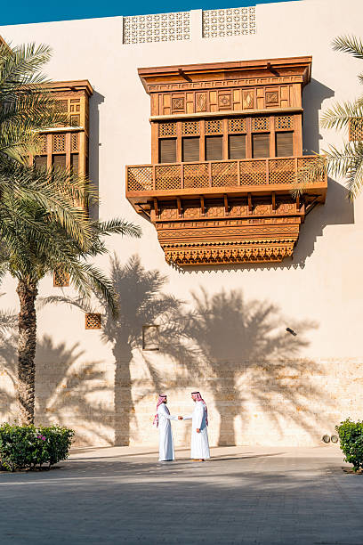 Two Arab men wearing traditional white thobes converse near a historic wooden balcony in Jeddah Cultural Square, surrounded by palm trees under a sunny blue sky.