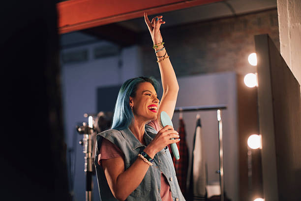 A vibrant singer with turquoise hair belts out a song, hair brush in hand, raising her arm in celebration; Backstage lights and wardrobe racks hint at a dynamic, casual performance moment