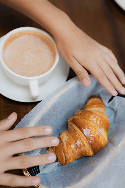Child's hands reaching for a fresh baked croissant in a basket next to a cup of hot chocolate on a wooden table