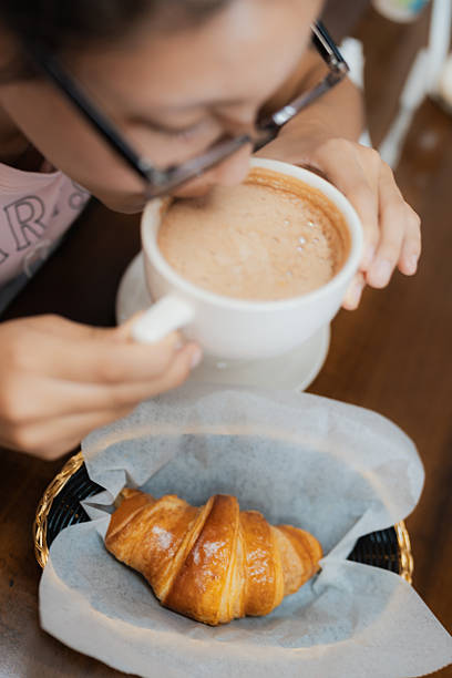 Young adult woman wearing glasses enjoying a warm beverage and a fresh croissant for breakfast or a coffee break