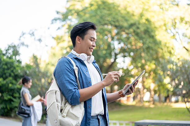 istock Asian man student carrying bag holding pen looking at tablet while standing or walking in university 2249657320
