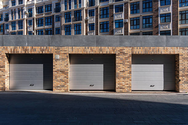 A modern brick garage with three individual sectional doors in grey. There are geometric shadows from the side sunlight, and an urban apartment building is visible in the background.