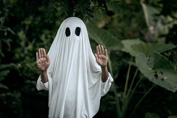 istock A young boy dressed as a ghost for Halloween wears a white sheet with two black eye holes. He raises both arms in a spooky gesture, surrounded by dark green foliage 2249666211