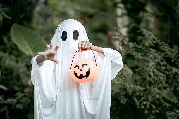 istock A young boy dressed as a ghost for Halloween wears a white sheet with black eye holes. Holding a pumpkin jack-oâ-lantern bucket, he stands outdoors surrounded by dark green foliage 2249666237