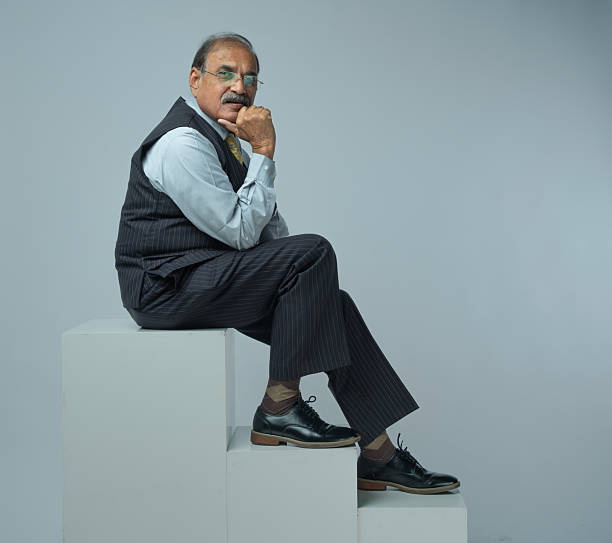 Full length of confident senior professional in suit and eyeglasses sitting on block with relaxed posture against white background