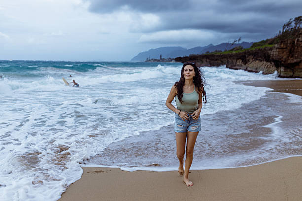 A young woman walks along a windy Hawaiian beach as strong waves roll in and surfers paddle behind her.