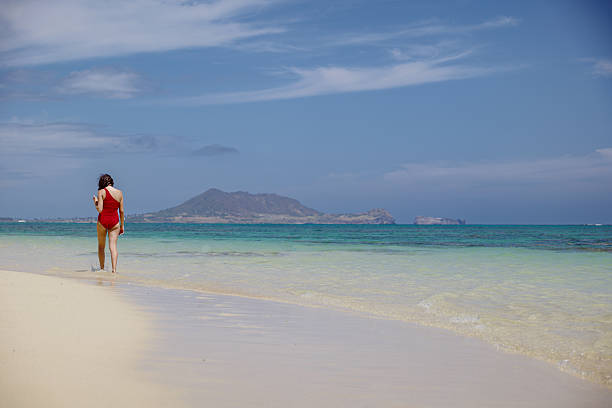 A young woman in a red swimsuit walks along the sandy shoreline at Lanikai Beach and enjoys the scenic views of the turquoise ocean and the Ulupa'u Head visible in the distance, while on vacation in Hawaii.