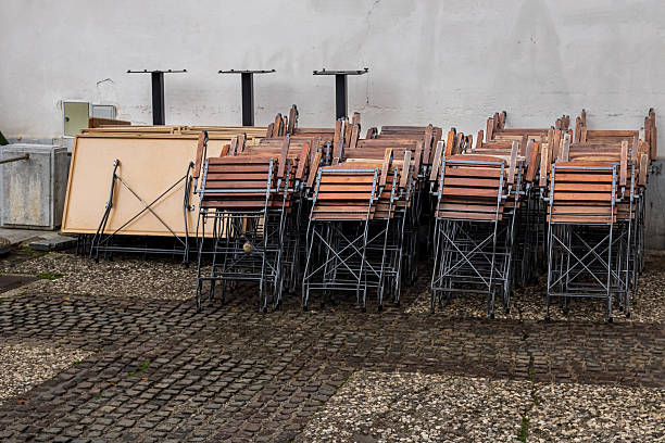 Wooden chairs stacked in rows and folded tables are stored outdoors against a wall. The chairs feature metal frames and are positioned on a cobblestone and concrete pavement. The tables are stacked vertically beside the chairs. The scene suggests a storage arrangement, possibly belonging to a nearby café or restaurant. The mix of stone and concrete ground provides a textured foundation beneath the stored furniture, while the white wall in the background offers a neutral backdrop.