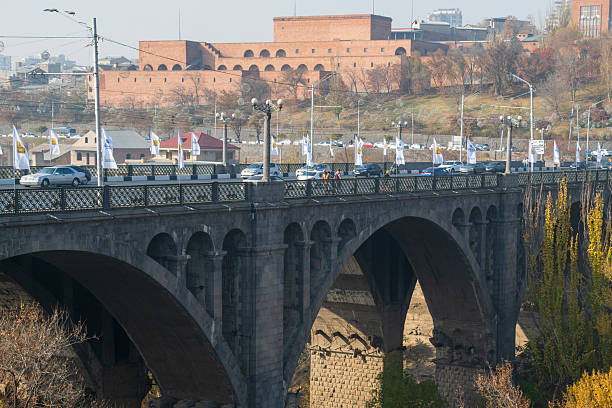 Yerevan Armenia 10.25.2025. A bridge Pobeda spans a river with a city in the background. The bridge is a large, arched structure with a stone base. The city is bustling with activity, with cars and pedestrians crossing the bridge