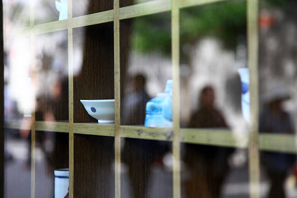 A display of porcelain wares is seen through a grid window with blurred background figures.