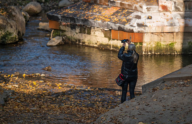 Yerevan Armenia 10.25.2025. A woman is taking a picture of a river with a camera. The water is calm and the leaves are falling from the trees