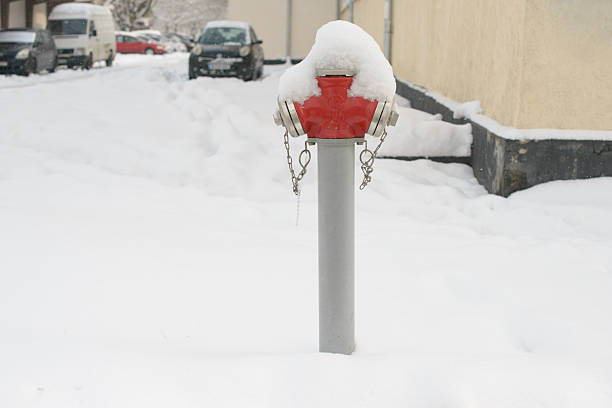 A red fire hydrant rises from deep snow in a quiet winter street, its metal caps and chains partly buried under fresh snowfall with parked cars and buildings in the background.