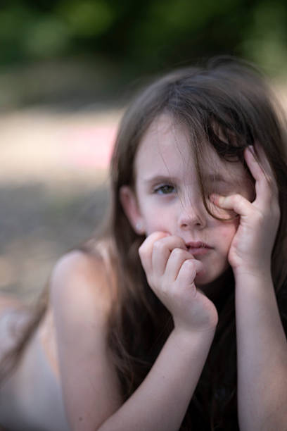 Squinting girl resting head on hands on beach, close up selective focus vertical summer portrait of relaxed childhood outdoor leisure with copy space