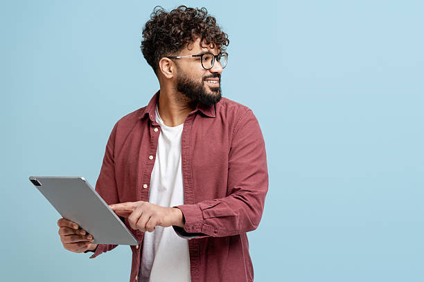 Young adult man smiling, using digital tablet, looking away on blue background
