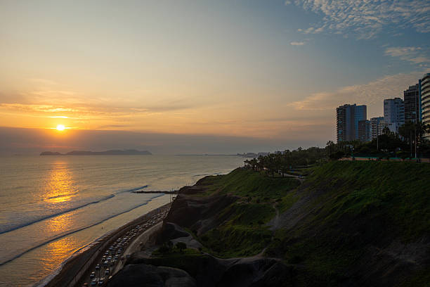 The sun sets over Isla San Lorenzo, casting a golden glow on the Pacific Ocean and the green cliffs of Lima, Peru. The serene coastal scenery offers a breathtaking view of nature’s beauty and tranquility
